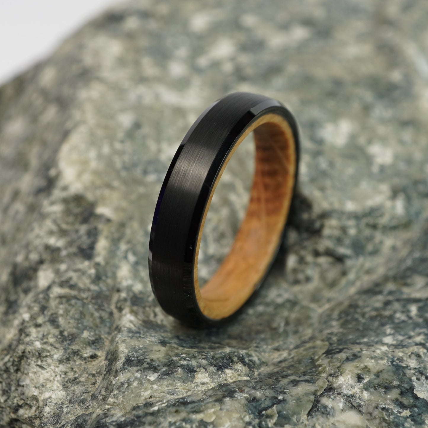 Black and barrel-wood bevelled ring, 4mm width, by Forge and Lumber, displayed on a textured stone surface.