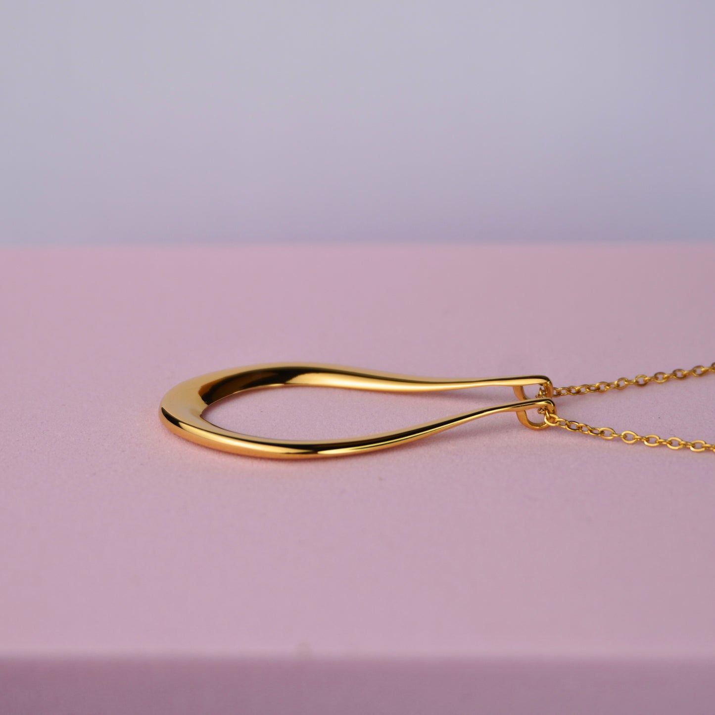 Gold teardrop pendant necklace on a delicate chain, displayed on a pink background.