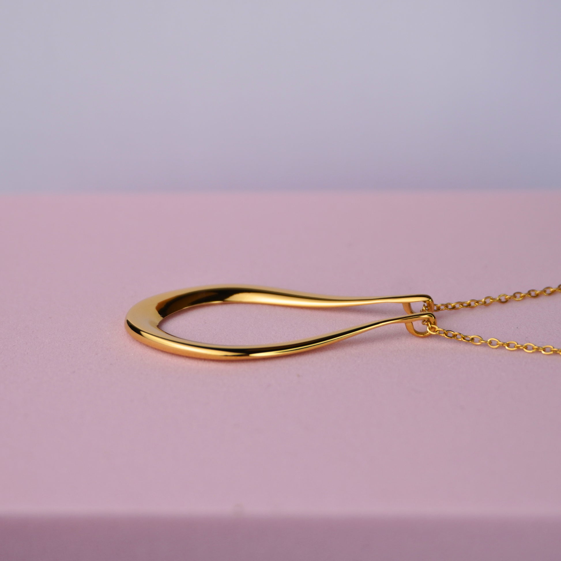 Gold teardrop pendant necklace on a delicate chain, displayed on a pink background.